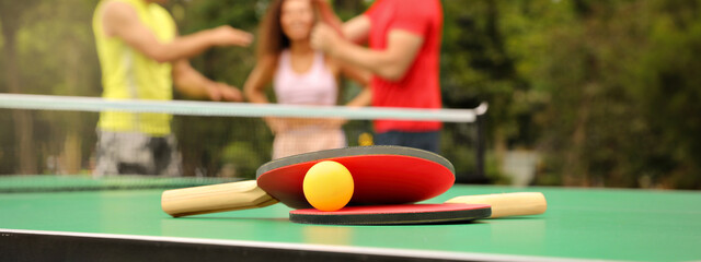 Friends talking near ping pong table outdoors, focus on rackets and ball. Banner design - obrazy, fototapety, plakaty