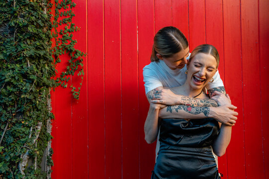 lesbian couple whisper in ear and laughing in front of red wall 