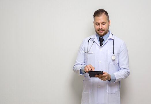Young caucasian doctor with short hair, mustache and beard. Dressed  in white gown and stethoscope typing on tablet computer. Stand in front of white wall.