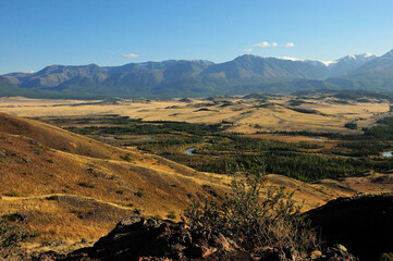 Stunning view from the top of the hill to the autumn steppe with a winding river flowing through it and snow-capped mountains in the distance.