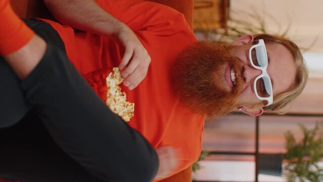 Portrait of redhead excited young man sits on couch eating popcorn and watching interesting tv serial, sport game, film, online social media movie content at home. Guy enjoying domestic entertainment