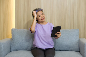 Asian retired elderly with eyeglasses, sitting on the sofa watching at tablet computer. With...