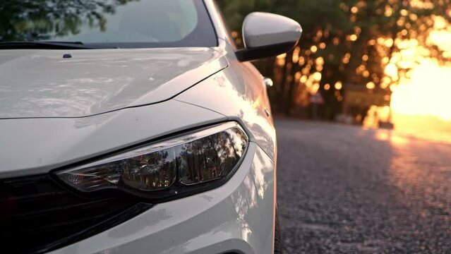 Close up view of front light of car headlights. unrecognizable white car is parked on side of road with setting sun in background. detail of exterior of modern car.