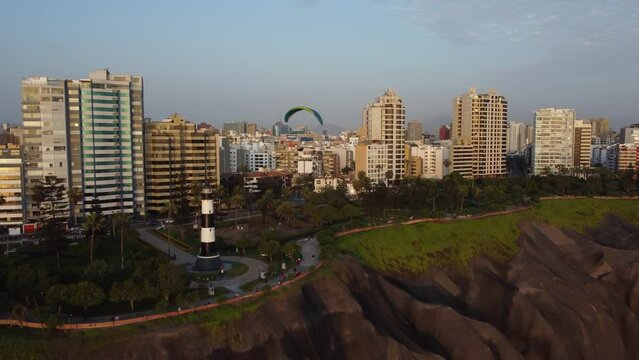 Drone 4k footage of lighthouse called "Faro de la Marina" in Miraflores district of Lima Peru. A paraglider flies above it. Drone orbits up revealing the buildings and the cityscape in the horizon.