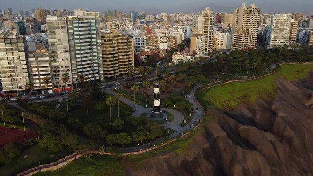 Drone 4k footage of a public park with a lighthouse called "Faro de la Marina" near a cliff's edge. Many trees surround it. Drone flies down while tilting camera up. Located in Lima Peru Miraflores.