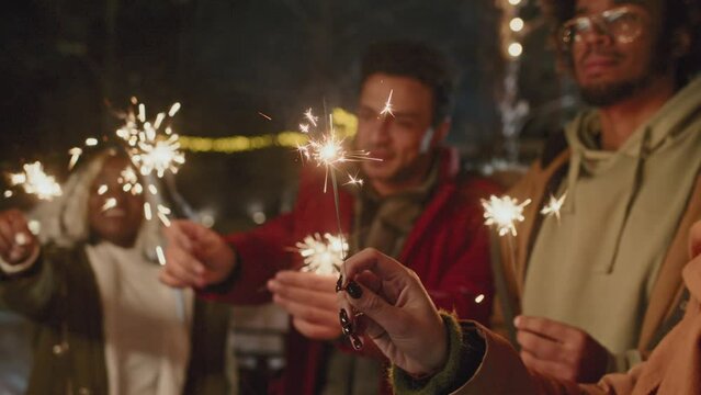 Selective focus of ethnically diverse young friends enjoying New Year party outdoors dancing with sparklers in hands