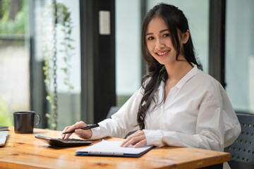 Confident beautiful Asian businesswoman using a calculator and taking notes in the office. Looking at the camera.