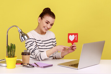 Smiling optimistic woman sitting at workplace, asking to rate her posts showing likes counter template to laptop screen. Indoor studio studio shot isolated on yellow background.