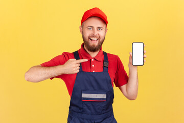 Portrait of joyful repairman in blue overalls red t-shirt pointing finger at smartphone with empty...