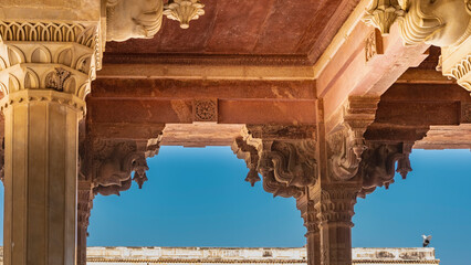 Details of ancient Indian architecture. The ceiling and columns with capitals and carved openwork stone ornaments are orange-pink. Blue sky. Jaipur. Amber Fort