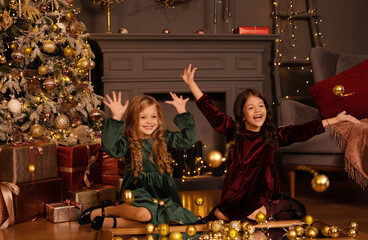 Two sisters playing near the Christmas tree and gifts. Happy little girls decorating christmas tree at home.