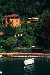 Scenic view of the white sailboat anchored near the shore of Varenna, Lombardy, Italy, with Italian villas overlooking the water of Lake Como, and botanical garden along the shore.