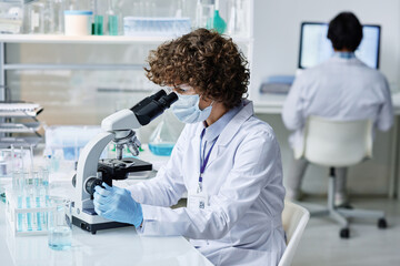 Young woman in labcoat, protective mask and gloves looking in microscope by workplace while carrying out scientific investigation