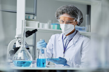 Young female researcher in protective workwear making clinical experiment while mixing two liquid...