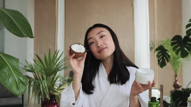 Blogger or content creator asian woman preparing natural cosmetics at home holding coconut homemade cream in glassware for skin and hair care home spa sitting at wooden table with exotic background
