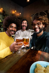 Vertical selfie of three male friends toasting happily in a bar, looking at camera with big smiles.