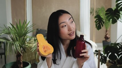 Beautiful healthy asian woman in white bathrobe sitting at wooden table with various ingredients preparing natural cosmetics at home, holding pumpkin and bottle with body milk or cream. Skin care.