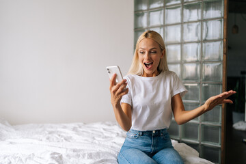 Portrait of millennial blonde young woman looking mobile phone laughing with good news or discount voucher for shopping at home. Cheerful female looking on cellphone app read message feel excited.