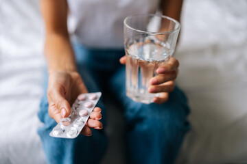 Close-up high-angle view of unrecognizable young woman holding pill in hand with water. Female going to take tablet from headache, painkiller, medication drinking clear water from glass, cropped shot.