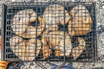 Overhead view of grilled slices of swordfish in a barbecue grill with a bread and blurred beach pebbles on background.