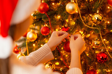 Close up back shot of a girl decorating Christmas tree with red and golden baubles. Beautiful artificial Christmas tree fully decorated in room