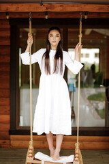 A cute brunette girl with long hair in a white dress poses while standing on a swing