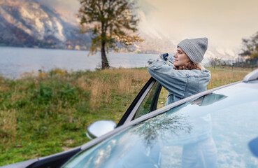 Young smiling caucasian woman driver traveler standing by the car admiring the view of the mountain lake