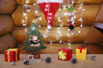 On a burlap-covered table are Christmas decorations and pine cones on the background of a garland and Santa hat hanging on a log wall.