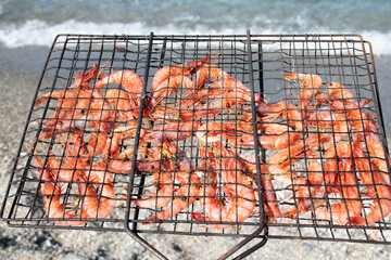 Grilled shrimp in a barbecue grill held in front of the sea with blurred beach pebbles and a wave on background