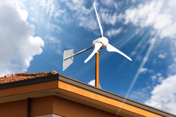 Close-up of a small wind turbine on the top of a roof of a house, against a blue sky with clouds...