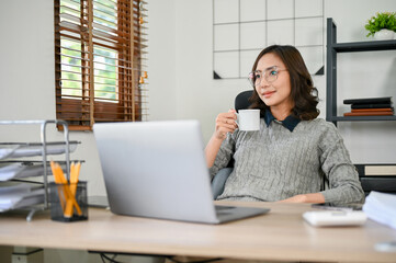 Successful Asian businesswoman or female boss sipping morning coffee at her desk in the office.
