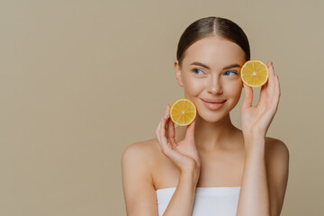 Horizontal shot of thoughtful young European woman uses homemade fruit for facial mask holds lemon slices wrapped in white soft bath towel looks aside isolated over brown background copy space