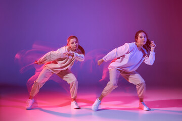 Fototapeta na wymiar Portrait of two young girls dancing hip-hop isolated over gradient violet background in neon with mixed light