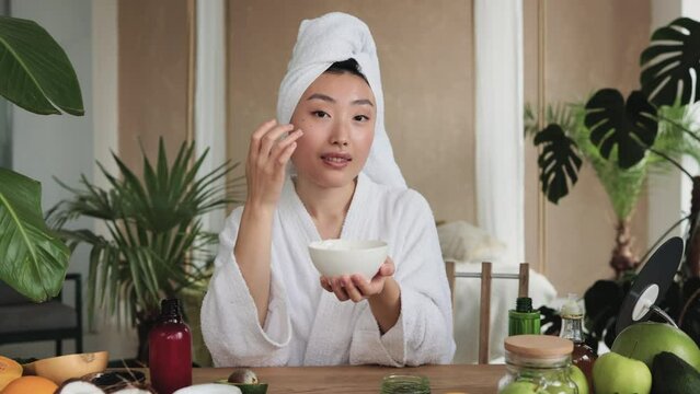 Beautiful asian woman in towel preparing homemade cream with natural ingredients for skin care applying on face sitting at wooden table with various organic fresh vegetables and fruits.