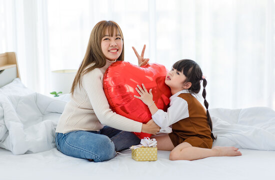 Millennial Asian young beautiful mother sitting on bed smiling holding hugging red heart shaped helium air balloon and present box with little cute preschooler daughter girl in bedroom in morning