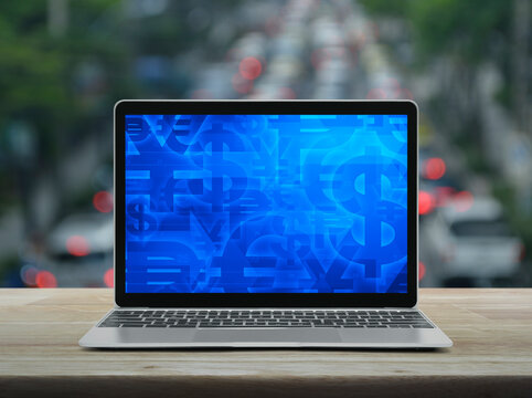 Financial currency symbol on modern laptop computer screen on wooden table over blur of rush hour with cars and road in city