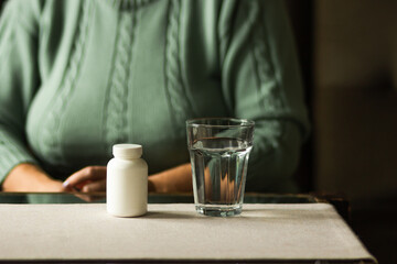 White plastic bottle with pills and glass of water on the table. Senior woman ready to take supplement pill. Cold winter