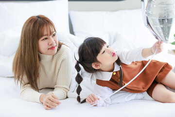 Asian young female teenager mother nanny babysitter sitting under thick warm white blanket smiling holding air balloons together with little cute preschooler daughter girl on bed in bedroom at home