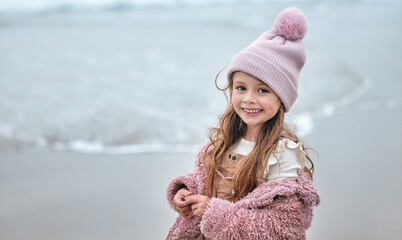 Happy girl child on beach, portrait on winter holiday with pink beanie and kid smile on the Dublin seaside. Outdoor freedom on ocean break, cute toddler relaxing by the water and coastal peace