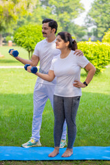 Indian trainer help a woman with dumbbell workout at park, working on arms strength, Asian athletic lady exercising outdoor with coach in nature, fitness concept.