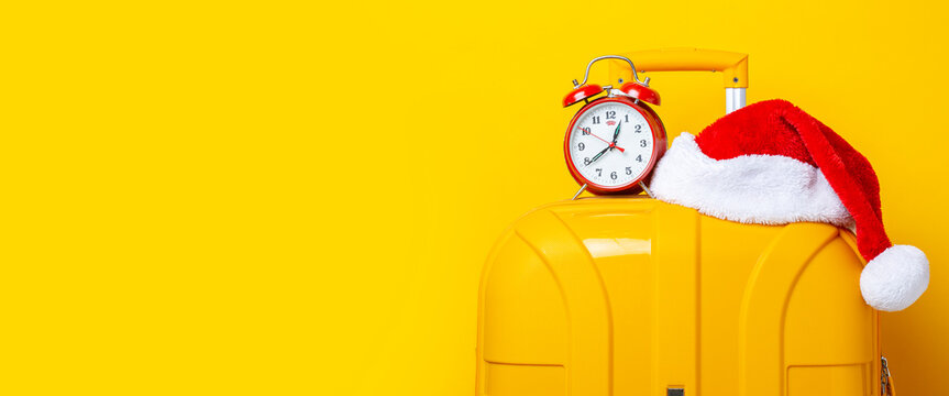 Alarm clock and Santa Claus hat lie on top of a yellow suitcase on a yellow background. Banner