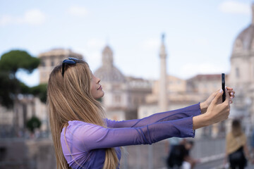 A young woman holds her phone while taking a selfie outside in Rome during the summer