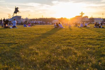 People with families relax on a green lawn in the evening at sunset near the Monument to Peter 1 on Senate Square in St. Petersburg. Abstract blurred background.