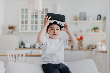 Little serious  boy taking of vr glasses, plays using virtual reality googles sitting on sofa over...