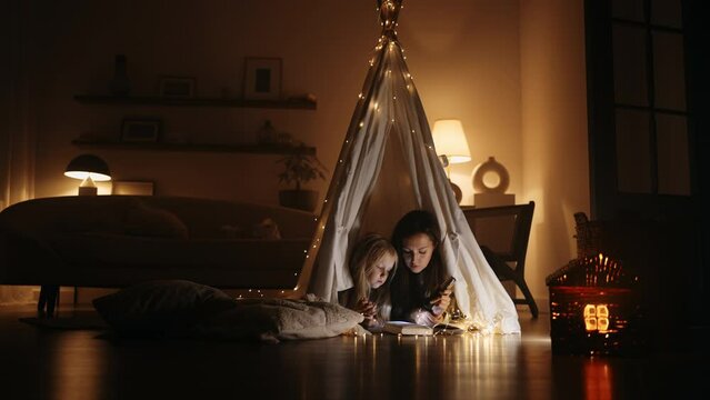 happy little girl and her mom are playing in teepee tent in room in evening, reading book