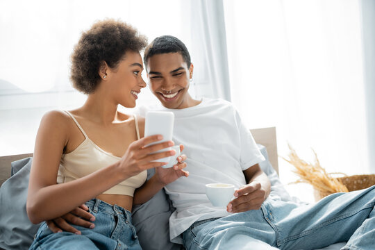 happy african american couple with coffee cups looking at smartphone on bed.