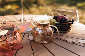 Cozy autumn picnic in the park. Close up of glass kettle with hot black tea, classic cups, homemade apple pie, maple leaves and burning candles on wooden table outdoors.