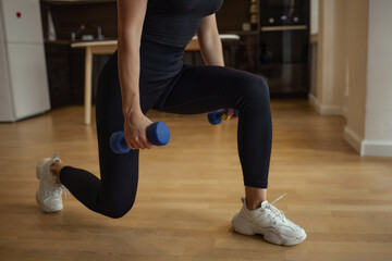 Naklejka premium The girl is engaged in fitness and trains her hands to lift dumbbells. the concept of a healthy lifestyle and activities at home. athletic blonde in a black suit