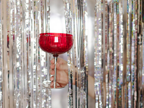 Woman's hand holding red glasses on the silver tinsel foil fringe background. Christmas celebration toast concept greeting card - Powered by Adobe