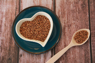 Small grains of natural brown buckwheat in a decorative plate in the form of a heart and in a wooden spoon